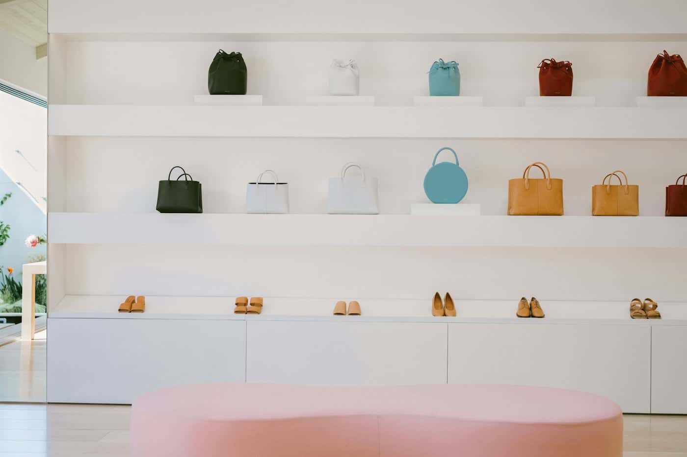 Leather handbags and shoes on backlit white shelves above a pink upholstered bench in a bright boutique