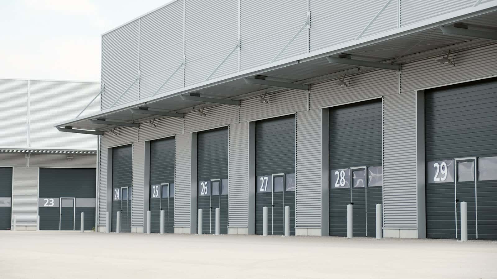 Grey metal-clad logistics building with a row of numbered dark sectional dock doors under a steel canopy
