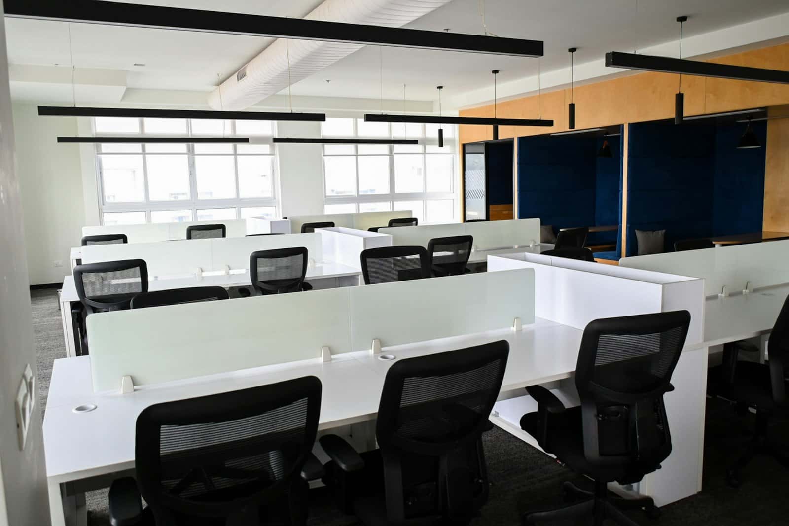 Rows of white desks with frosted screens and black mesh chairs, with navy upholstered booths along a timber-clad wall