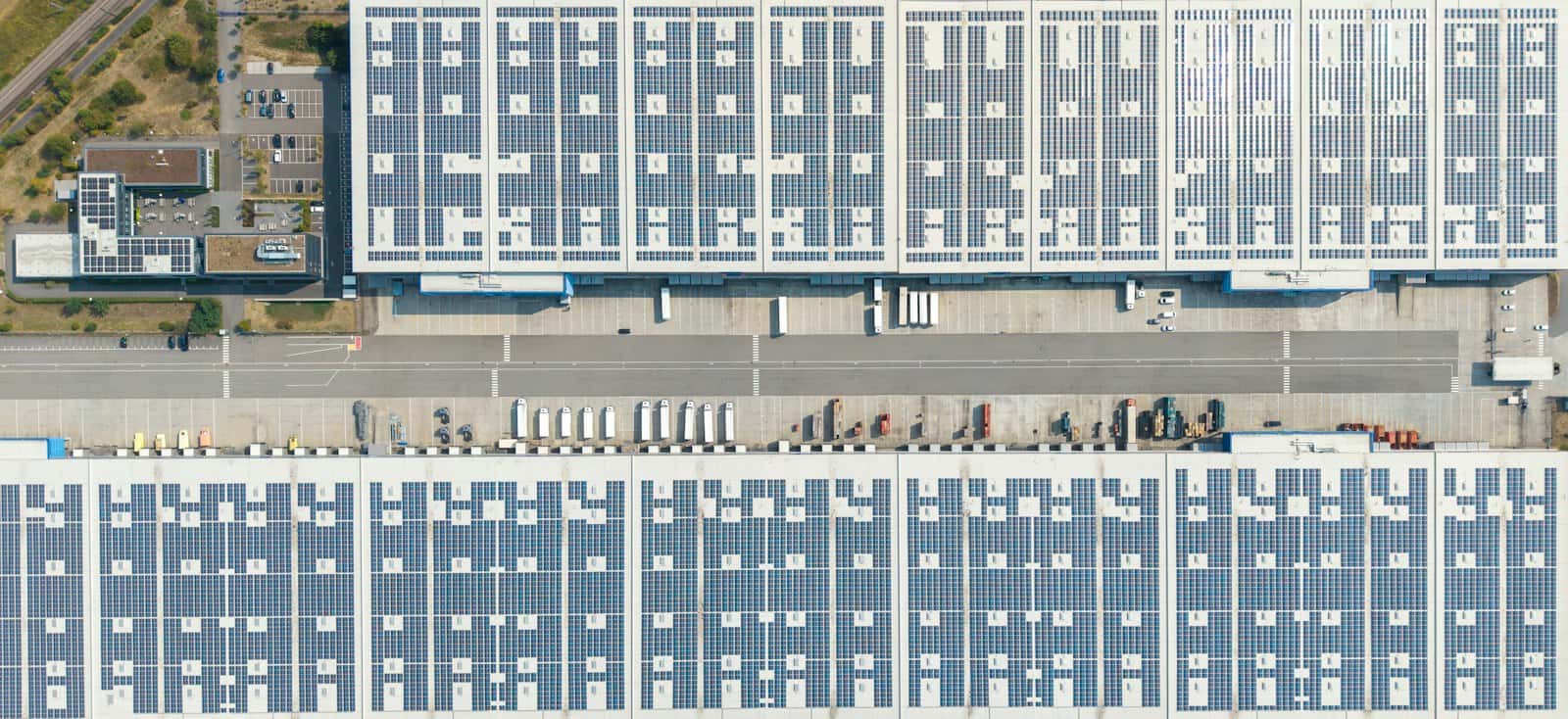 Overhead aerial of two long warehouse roofs covered in solar panels, facing a shared truck court with trailers at the docks