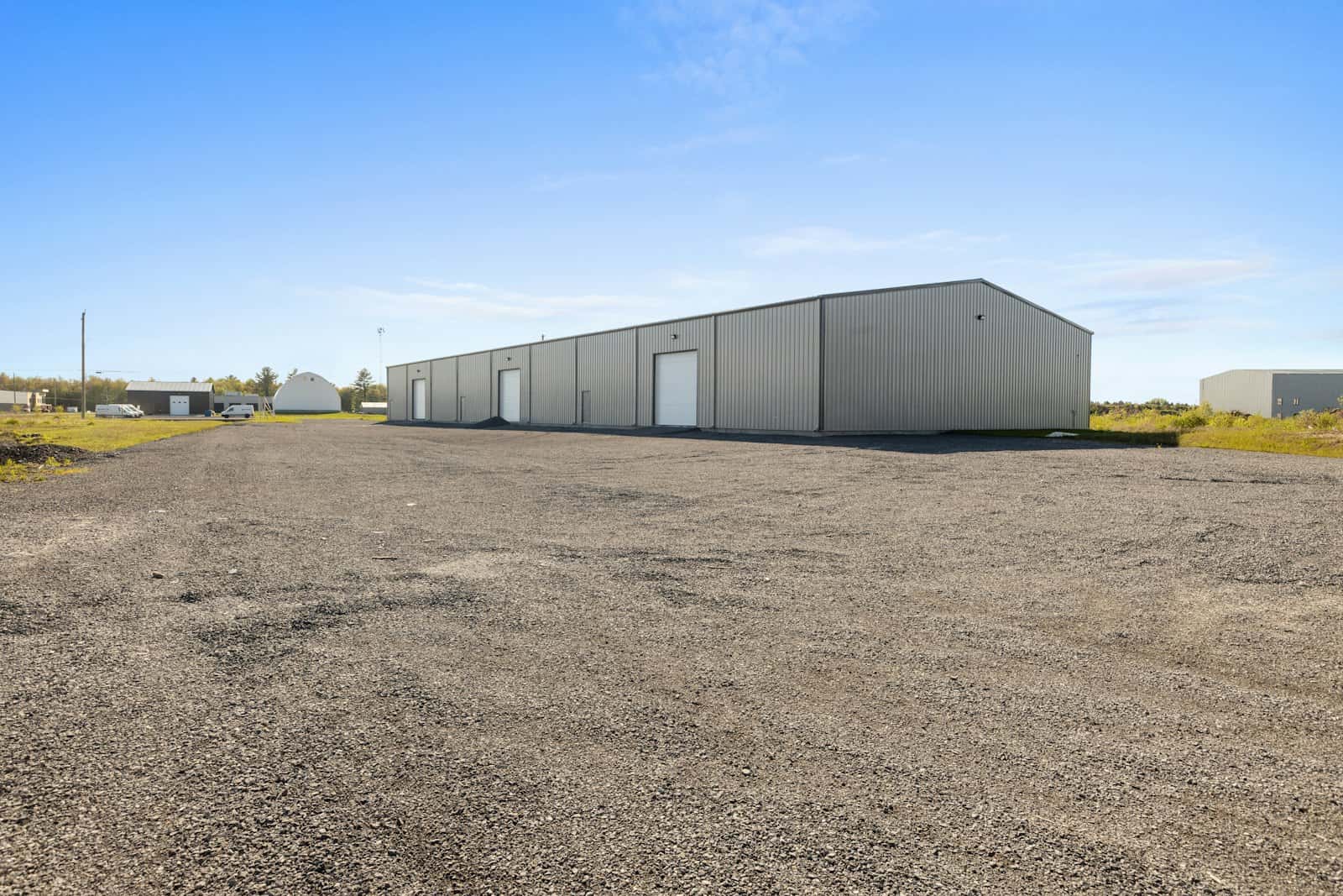 Long pre-engineered metal shed with roller doors along its side, set behind a wide gravel yard under a clear sky