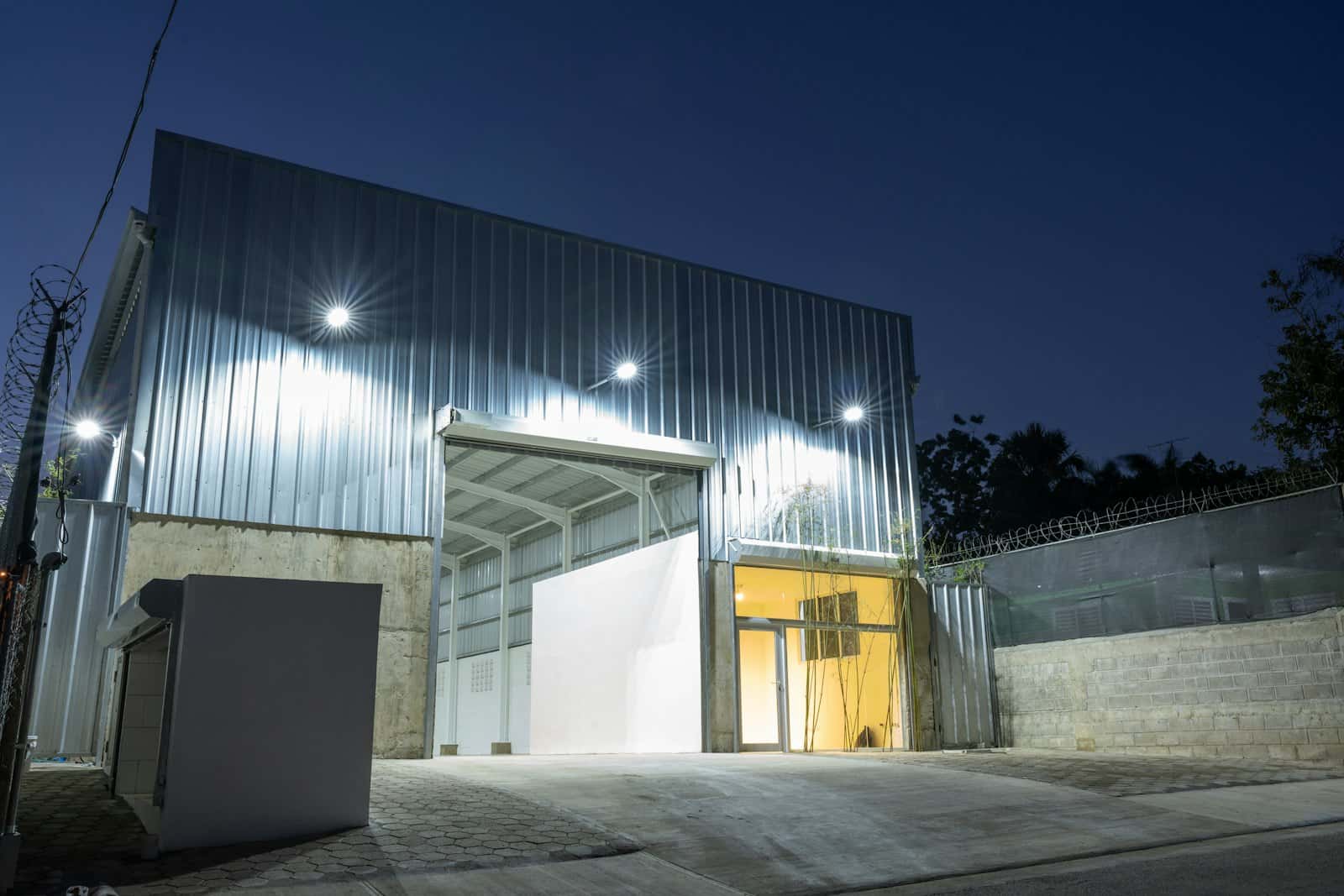 Compact metal-clad industrial building lit at dusk, with a tall open loading bay and a glazed office entrance